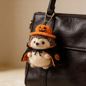 Könnte beinhalten: Ein Halloween-Hedgehog-Schlüsselanhänger. Der Plüsch-Igel trägt einen schwarzen Umhang mit orangefarbenem Futter und einen Kürbishut. Der Schlüsselanhänger ist an einer dunkelbraunen Ledertasche befestigt.
