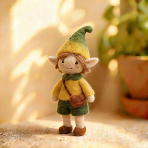 Könnte beinhalten: Eine handgefertigte Elfenpuppe aus Filz mit gelb-grünem Hut und passendem Hemd. Die Puppe hat braune Shorts, eine braune Umhängetasche und braune Schuhe. Der Elf hat spitze Ohren, braune Haare und ein lächelndes Gesicht.