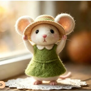 Könnte beinhalten: Eine handgefertigte Filzmaus-Figur, die ein grünes Kleid und einen Strohhut trägt. Die Maus hat große Ohren, eine rosa Nase und steht auf einer weißen Spitzendecke. Die Figur ist ein charmantes Beispiel für handwerkliche Arbeiten.