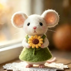 Könnte beinhalten: Eine handgefertigte weiße Mausfigur mit großen Ohren, die ein grünes Kleid trägt und eine gelbe Sonnenblume hält. Die Maus hat rosa Ohren, Nase und Füße. Die Figur steht auf einer weißen Spitzendecke.