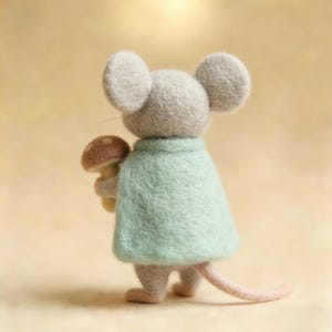 May include: A felt mouse figure, seen from behind, wearing a pale green cape and clutching a mushroom. The mouse is grey with large, round ears and a pink tail. The mushroom has a brown cap and a yellow stem.