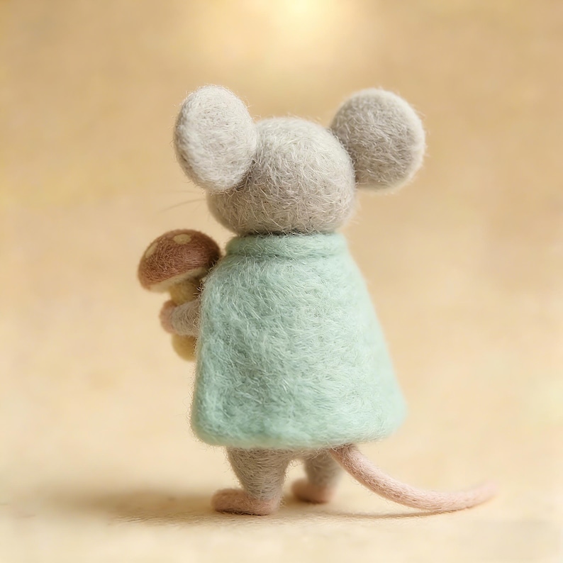 May include: A felt mouse figure, seen from behind, wearing a pale green cape and clutching a mushroom. The mouse is grey with large, round ears and a pink tail. The mushroom has a brown cap and a yellow stem.