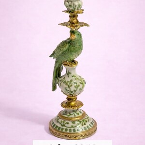May include: A candlestick holder featuring a green parrot perched on a white ceramic base with green vine patterns. The structure includes gold-toned metal accents at the base, middle, and top candle cup, which also features a green vine design.