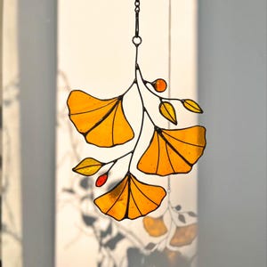 Puede incluir: Un atrapasoles de vidrieras con tres hojas de ginkgo de color amarillo vibrante sobre una rama de metal oscuro. La pieza incluye pequeños detalles en naranja y amarillo y está suspendida por una fina cadena de metal, creando un adorno botánico.