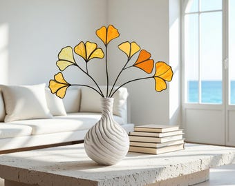 May include: A white, textured vase with a swirling design holds a decorative arrangement of yellow and orange stained glass ginkgo leaves. The vase sits on a white stone coffee table, with a stack of books nearby.
