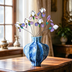 Op de afbeelding: Een blauwe keramische vaas met een textuurrijke, gevlekte afwerking bevat een verzameling irissen van glas-in-lood. De bloemen hebben witte en paarse bloemblaadjes met groene accenten, elk bevestigd aan een dunne, donkere metalen stengel.