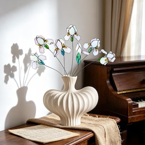 May include: A white ceramic vase with a ribbed, rounded body holds a bouquet of stained glass iris flowers. The flowers feature white petals with yellow and green accents, connected by thin dark metal stems, creating a delicate floral display.