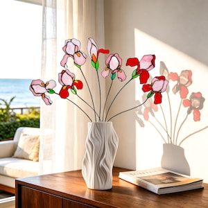 May include: A white ceramic vase with a vertical wavy texture holds a bouquet of stained glass iris flowers. The flowers feature light pink and deep red petals with green stems and black outlines, creating a vibrant display of floral art.