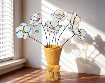 May include: A bouquet of six white stained glass flowers with yellow centers and thin black stems, arranged in a matte yellow vase shaped like a wrapped paper bouquet with a decorative bow tied around the center of the base.