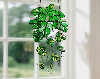 May include: Stained glass hanging plant featuring vibrant green Monstera leaves. The leaves have a detailed design with a mix of light and dark green hues, and are outlined in black. The plant is suspended by a chain, creating a decorative window accent.
