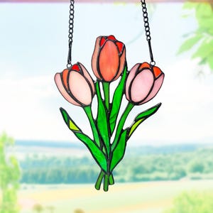 以下が含まれることがあります： ピンクとオレンジ色の3つの花が特徴のステンドグラスのチューリップの花束。緑の葉がアクセントになっています。黒いチェーンで吊り下げられており、窓のディスプレイに最適です。背景には美しい景色が広がっています。