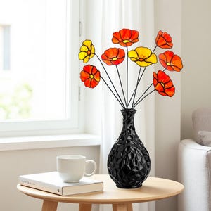 May include: A black textured vase holds a bouquet of stylized flowers with red and yellow petals. The vase sits on a wooden table next to a white mug and a book. The flowers have black stems.