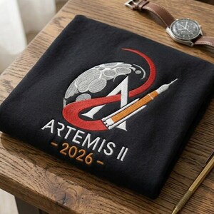 May include: Black square-folded Artemis II t-shirt with embroidered design. The design includes a stylized moon, a red curved line, the letter "A", and a rocket. The text "ARTEMIS II - 2026 -" is embroidered in white. A brown leather watch and a gold pen are on the wooden surface.