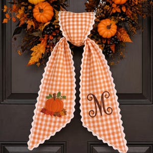 May include: A decorative fabric door hanger featuring an orange and white gingham pattern with scalloped edges. One side displays an embroidered orange pumpkin with green leaves, while the other side features a large, brown embroidered letter M.