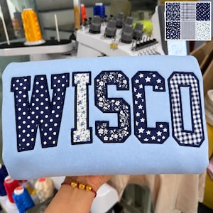 May include: A light blue sweatshirt featuring the letters WISCO in large, block-style applique. Each letter is filled with a different navy blue and white pattern, including polka dots, floral prints, stars, and gingham checks, with dark borders.
