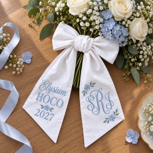 May include: A white fabric bow with long tails featuring blue embroidery. The left tail reads Elysium HOCO 2027 with small blue flowers. The right tail features a blue monogram of the letters RJ surrounded by blue floral embroidery.