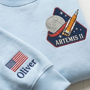 May include: Light blue sweatshirt with embroidered patches. One patch features a rocket, moon, and the text "ARTEMIS II." The other sleeve has an American flag patch and the name "Oliver" embroidered in navy blue.