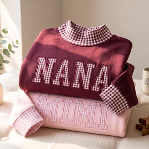 May include: A maroon sweatshirt with a high collar and long sleeves featuring a white and maroon gingham pattern, with the word NANA embroidered in gingham. Beneath it, a light pink sweatshirt with the word MIMI embroidered in light pink.