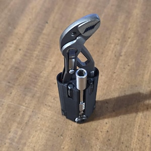 May include: A multi-tool with a silver adjustable wrench and a black base containing a socket wrench. The tool is designed for various repair tasks. The image is taken from a slightly elevated angle.