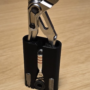 May include: A multi-tool with a silver metal adjustable wrench and other tools, housed in a black rectangular case. The wrench is open, revealing its serrated jaws. The tool is set against a light brown wooden surface.