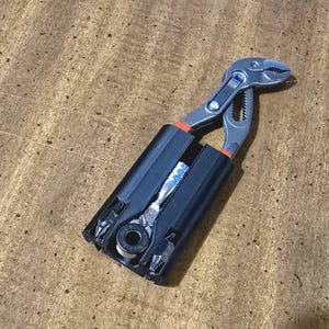 May include: A multi-tool kit featuring a silver and black adjustable wrench and a small ratchet with various bits. The tools are housed in a black case with orange accents, set against a wood grain surface.