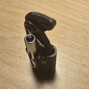 May include: A black multi-tool with a silver socket wrench head. The tool is compact and designed for various mechanical tasks. It is resting on a light brown wooden surface.