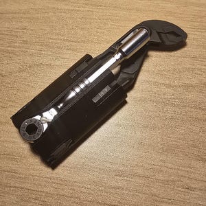 May include: A black multi-tool with a silver ratchet and pliers. The tool is partially open, revealing the ratchet and a socket. The tool is on a wooden surface.