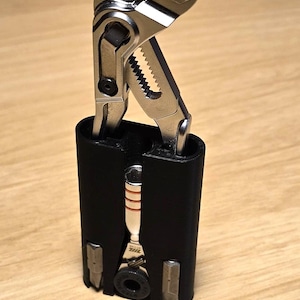 May include: A multi-tool with a silver metal pliers head and a black handle. The tool is partially inserted into a black case. The case has several other tools attached to it. The background is a light brown wooden surface.