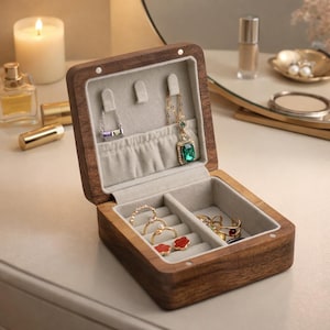 May include: A wooden jewelry box with a hinged lid, open to reveal a gray velvet interior. The box contains rings, necklaces, and earrings. The exterior is a dark brown wood. A lit candle and perfume bottle are in the background.
