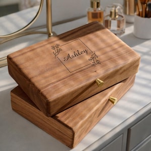 May include: A wooden jewellery box with a rectangular shape and a natural wood grain finish. The box features a gold-coloured knob and the name "Ashley" engraved within a floral frame on the lid. The box is stacked on top of another box.