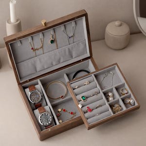 May include: Open wooden jewellery box with multiple compartments, lined with grey fabric. The box contains watches, bracelets, necklaces, and rings. The box is brown with a gold-coloured clasp.