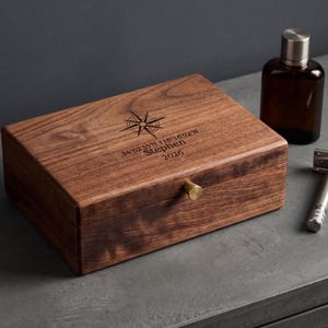 May include: A rectangular dark wood box featuring a brass knob and an engraved compass rose design with the text 34°02'35"N 118°16'02"W, the name Stephen, and the year 2026. A brown glass bottle and a silver metal safety razor are positioned nearby.