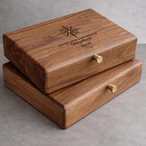 May include: Two stacked, rectangular wooden boxes with brass knobs. The top box features a compass rose design and engraved text: "34°02'35"N 118°16'02"W Stephen 2026". The boxes are a warm brown color.