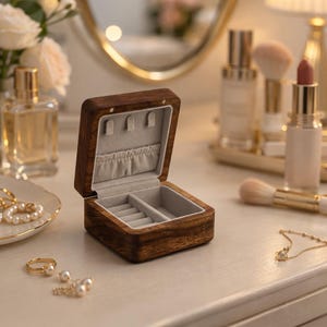 May include: Open wooden jewelry box with a dark brown finish and a light gray interior. The box is rectangular with a hinged lid and compartments for rings and other small jewelry items. The box is sitting on a white surface with other beauty products.