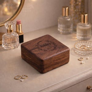 May include: A square, wooden jewelry box with a dark brown finish. The lid is engraved with the name "Ashley" inside a floral wreath and the year "2026". The box is on a light-colored surface, with perfume bottles and rings nearby.
