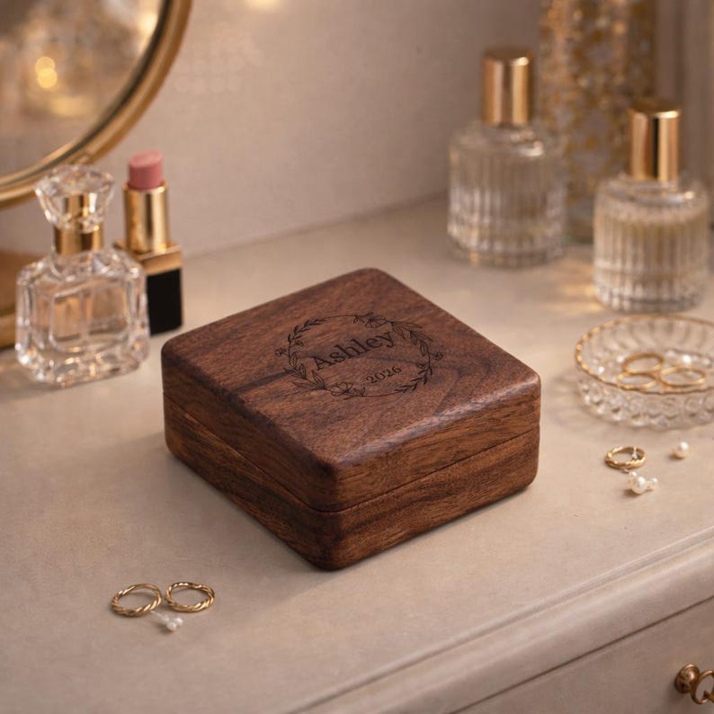 May include: A square, wooden jewelry box with a dark brown finish. The lid is engraved with the name "Ashley" inside a floral wreath and the year "2026". The box is on a light-colored surface, with perfume bottles and rings nearby.