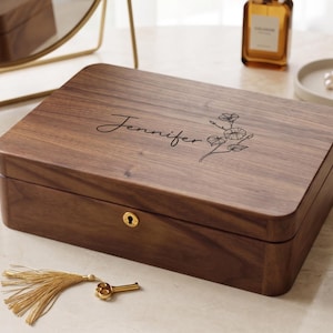 May include: A rectangular dark brown wooden jewellery box with a hinged lid featuring the engraved name Jennifer and a floral line drawing. The front includes a gold-coloured keyhole, and a gold-coloured key with a matching tassel lies nearby.