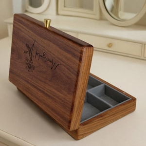 May include: A wooden jewellery box with a hinged lid, open to reveal a grey felt-lined interior with compartments. The box is engraved with the word "Marigold" and a floral design. A small gold-coloured knob serves as the handle.