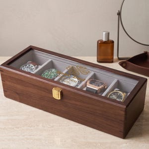 May include: A wooden watch box with a clear glass top, containing several wristwatches with various colored watch faces. The box has a gold-coloured clasp and is sitting on a light-coloured surface.