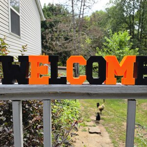 May include: A three-dimensional sign spelling the word WELCOME in alternating black and bright orange block letters. The letters are arranged in a single horizontal row, featuring a bold, sans-serif font style with a flat base for stability.