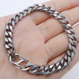 May include: A silver-colored curb chain bracelet with a secure clasp. The bracelet is made of interlocking links and is displayed on a light background.