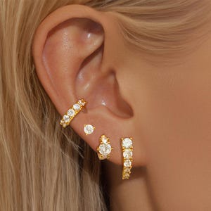 Könnte beinhalten: Goldfarbenes Ohrschmuck-Set mit verschiedenen Stilen. Enthält einen gebogenen Ohrklemme, Ohrstecker und kleine Creolen, alle mit klaren, funkelnden Steinen besetzt. Der Schmuck wird an einem Ohr präsentiert.