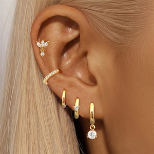May include: An ear displaying a collection of gold earrings. There's a cluster stud, a curved ear cuff, and three hoop earrings, two with small clear stones and one with a dangling round stone. The jewellery is set against a background of blonde hair.