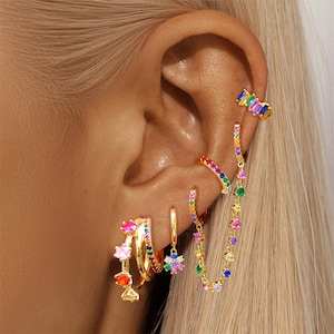 Könnte beinhalten: Eine Sammlung goldfarbener Ohrringe mit bunten Edelsteinen. Die Ohrringe umfassen Creolen, Ohrklemmen und Hängevarianten mit einer Vielzahl von Regenbogenfarbenen Steinen. Der Schmuck wird an einem Ohr präsentiert und zeigt die verschiedenen Designs und Farben.