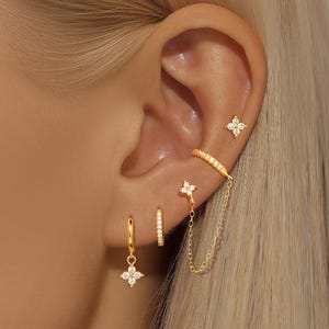 May include: A close-up of a person's ear adorned with multiple gold earrings. The earrings include small hoops, studs, and a chain earring, all featuring clear, sparkling gemstones and floral designs. The jewelry is set against the backdrop of blonde hair.
