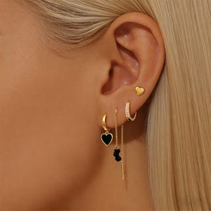 Puede incluir: Primer plano de una oreja adornada con múltiples pendientes dorados. Uno es un pendiente de corazón, otro es un aro pequeño con un colgante de corazón negro y un tercero es un aro pequeño con piedras transparentes. También se ve un pendiente de cadena con cuentas negras.