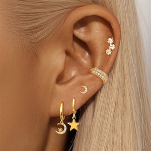 以下が含まれることがあります： ゴールドトーンの天体モチーフのイヤリングとイヤーカフ。イヤリングには、星が付いた三日月とぶら下がっている星が含まれています。小さな三日月スタッドとトリプルスターのスタッドも表示されます。イヤーカフには、小さな透明な石が飾られています。