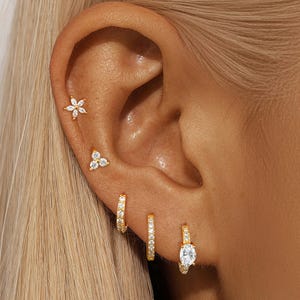 May include: Close-up of an ear adorned with multiple gold earrings. The earrings include a flower stud, a cluster stud, and three hoop earrings, two with clear stones and one with a single oval stone. The ear is set against a background of blonde hair.