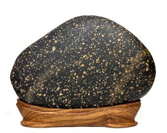 May include: A dark, speckled stone with a smooth, rounded shape, displayed on a carved wooden stand. The stone's surface is black with numerous small, golden-brown flecks. The wooden stand has a natural wood grain finish.