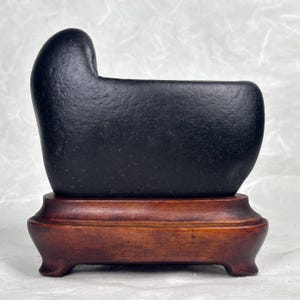 May include: A black stone sculpture with a rounded top and a rectangular body, set on a dark brown wooden stand. The stone has a textured surface. The sculpture is displayed against a neutral background.